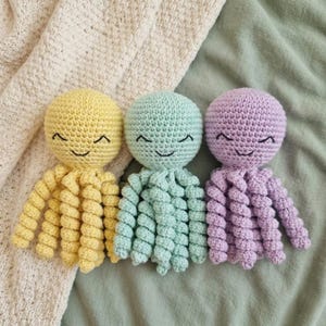 Pode incluir: Três brinquedos de polvo de crochê em cores pastel: amarelo, verde menta e lavanda. Cada polvo tem um rosto sorridente com olhos e boca bordados em preto. Os tentáculos são enrolados. Os brinquedos estão em uma superfície verde.