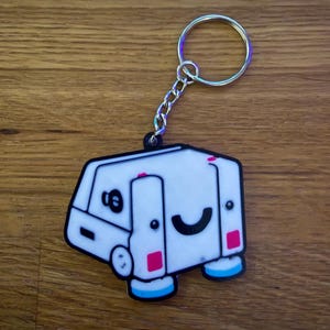 May include: A white camper keychain with a black outline, a smiling face, and pink and blue accents. The keychain has a silver chain and a silver ring.
