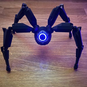 May include: A black, mechanical spider robot with eight articulated legs. The robot has a central body with a glowing blue circular light. The legs are positioned to support the robot on a wooden surface.