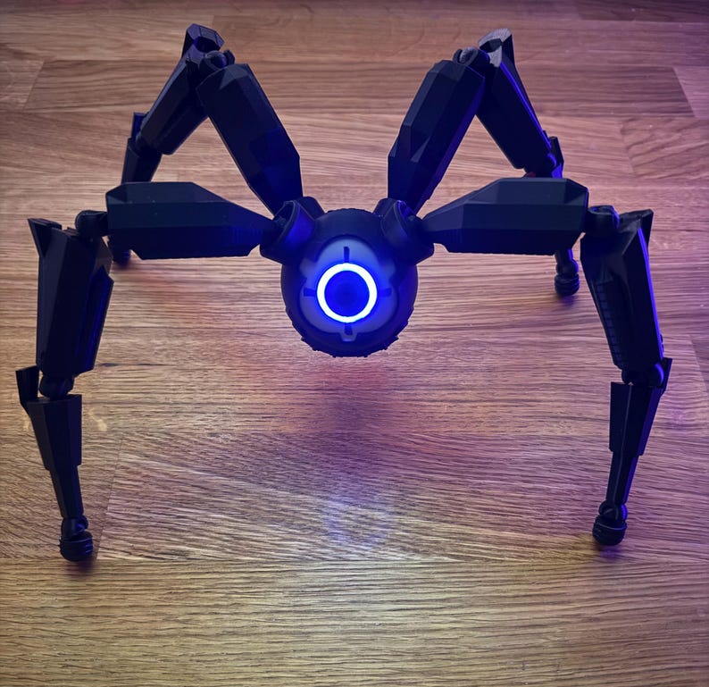 May include: A black, mechanical spider robot with eight articulated legs. The robot has a central body with a glowing blue circular light. The legs are positioned to support the robot on a wooden surface.