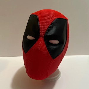 Könnte beinhalten: Eine rot-schwarze Deadpool-Maske. Die Maske hat einen strukturierten roten Stoff mit schwarzen Augen- und Mundbereichen. Die Augenöffnungen haben ein weißes Netzmaterial. Die Maske wird auf einem weißen Ständer präsentiert.