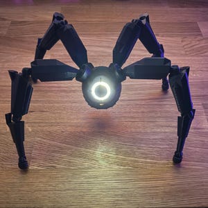 May include: A black, mechanical spider robot with eight articulated legs. The robot has a central body with a glowing white circular light. The legs are angled and positioned to support the robot.