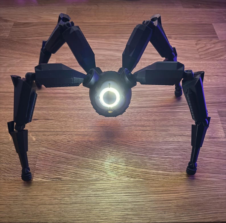 May include: A black, mechanical spider robot with eight articulated legs. The robot has a central body with a glowing white circular light. The legs are angled and positioned to support the robot.