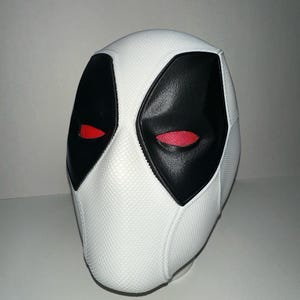 Op de afbeelding: Een wit masker met zwarte oog- en mondvormen en rode oogopeningen. Het masker heeft een textuuroppervlak en is waarschijnlijk een kostuum- of cosplay-item.