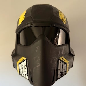 May include: A black helmet with a visor and yellow accents. The helmet has a rugged, worn appearance with visible scratches and paint splatters. The design includes rectangular yellow panels with white and black details.