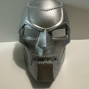 Può includere: Una maschera argentata metallica con un design dettagliato. La maschera presenta aperture rettangolari per gli occhi e una placca facciale angolare prominente. Piccoli rivetti distanziati adornano i bordi, aggiungendo consistenza e dettagli all'accessorio a tema supereroi.
