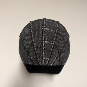 Op de afbeelding: Een zwart-grijze baseballcap met een textuur, webachtig patroon. De cap heeft een gebogen klep en een gestructureerde kroon. Het ontwerp heeft verticale en horizontale lijnen, waardoor een spinnenweb ontstaat.