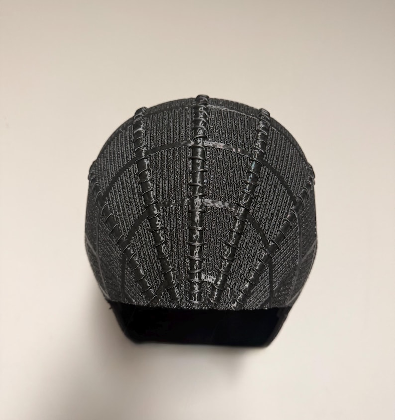 Op de afbeelding: Een zwart-grijze baseballcap met een textuur, webachtig patroon. De cap heeft een gebogen klep en een gestructureerde kroon. Het ontwerp heeft verticale en horizontale lijnen, waardoor een spinnenweb ontstaat.