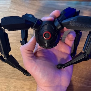 May include: A black, articulated, mechanical spider toy with multiple legs and a central body. The toy has a detailed design with a red circular element on the front. The toy is held in a hand.