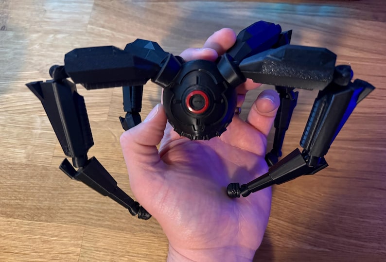May include: A black, articulated, mechanical spider toy with multiple legs and a central body. The toy has a detailed design with a red circular element on the front. The toy is held in a hand.