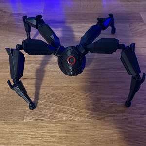 May include: A black, mechanical spider-like model with articulated legs and a central body. The body features a red circular detail. The model is positioned on a wooden surface, showcasing its intricate design and robotic aesthetic.
