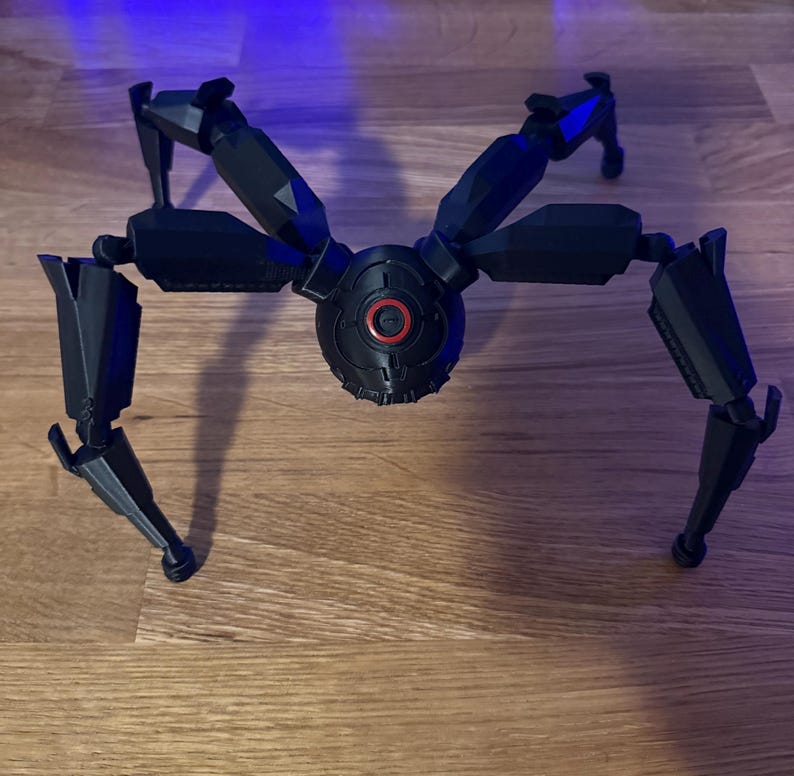 May include: A black, mechanical spider-like model with articulated legs and a central body. The body features a red circular detail. The model is positioned on a wooden surface, showcasing its intricate design and robotic aesthetic.