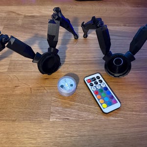May include: Black articulated robotic legs with circular bases, a small LED light, and a remote control with color options. The items are arranged on a wooden surface.