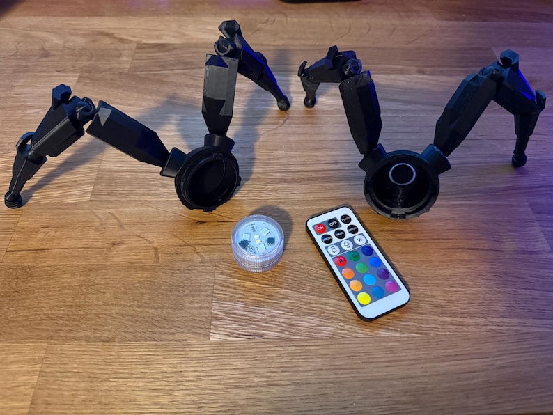 May include: Black articulated robotic legs with circular bases, a small LED light, and a remote control with color options. The items are arranged on a wooden surface.