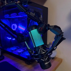 May include: A black and silver microphone with a blue and green illuminated honeycomb pattern. The microphone is held by a black articulated arm. The background includes a computer tower with blue lighting.