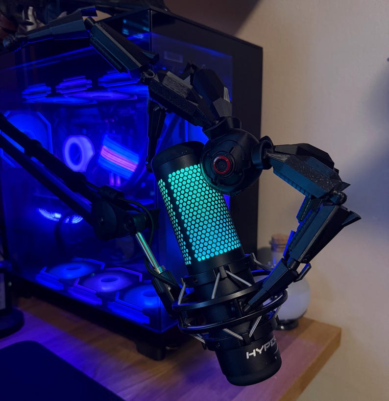 May include: A black and silver microphone with a blue and green illuminated honeycomb pattern. The microphone is held by a black articulated arm. The background includes a computer tower with blue lighting.
