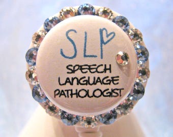 May include: A white badge with blue rhinestones around the edge. The badge says "SLP Speech Language Pathologist" with a heart drawn next to the letters "SLP".