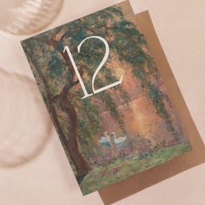 Puede incluir: Una tarjeta de felicitación con una pintura de una escena junto al agua con cisnes y el número 12 en blanco. La tarjeta está sobre un sobre marrón, con dos vasos de agua en la esquina superior izquierda.