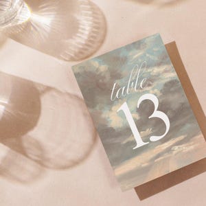 Könnte beinhalten: Eine Tischnummerkarte mit einem bemalten Himmelshintergrund. Die Nummer 13 ist in Weiß auf die Karte gedruckt. Der Text "Tisch" ist in Weiß über der Nummer gedruckt.