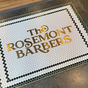 Könnte beinhalten: Ein weißer Fliesenboden mit grünem Rand und goldener Beschriftung, die "The Rosemont Barbers" lautet.
