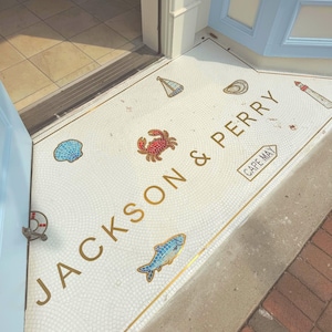 以下が含まれることがあります： 金色の文字でJACKSON & PERRYと書かれた白いモザイクタイルの床。ヨット、貝殻、赤いカニ、青い魚、灯台、CAPE MAYと書かれた看板のカラフルなタイルイラストがデザインされています。