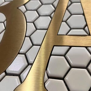 May include: Close-up of the letters "BA" in gold metal, mounted on a white hexagonal tile background.