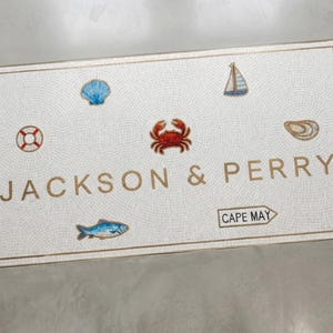 May include: White mosaic doormat with a gold border, featuring nautical illustrations of a crab, seashell, sailboat, and lighthouse. The text "JACKSON & PERRY" and "CAPE MAY" are displayed.