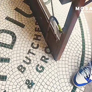 Puede incluir: Un suelo de baldosas de mosaico con las palabras "BUTCHERS & GRILL" en letras de color verde oscuro. El suelo es blanco con un patrón de cuadrícula y un borde verde. Una persona lleva zapatillas deportivas azules y blancas.