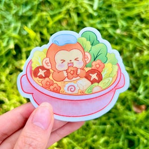 May include: A die-cut sticker featuring a cartoon monkey with a light blue towel on its head, sitting in a pink bowl of ramen. The bowl contains green leafy vegetables, sliced mushrooms, a white tofu cube, and a pink and white narutomaki swirl.