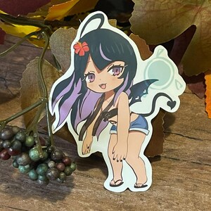 May include: A sticker of a cartoon character with black hair, purple streaks, and a red flower. The character wears a brown top and blue shorts, with black wings and a tail. A light blue sun-like shape is behind the character.