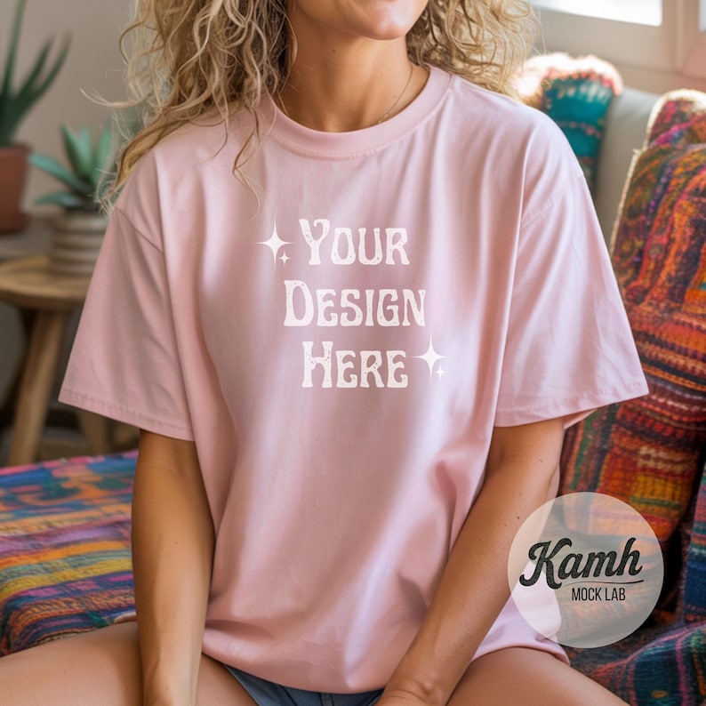 May include: A light pink t-shirt with the text "YOUR DESIGN HERE" in white, with star accents. The shirt is a mock-up, and the brand "Kamh MOCK LAB" is visible in the lower right corner.