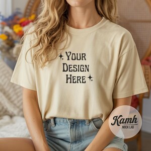 May include: Cream-colored t-shirt with the text "YOUR DESIGN HERE" in a black, vintage-style font. The shirt is a short-sleeved, crew neck style. The model is wearing blue denim shorts.