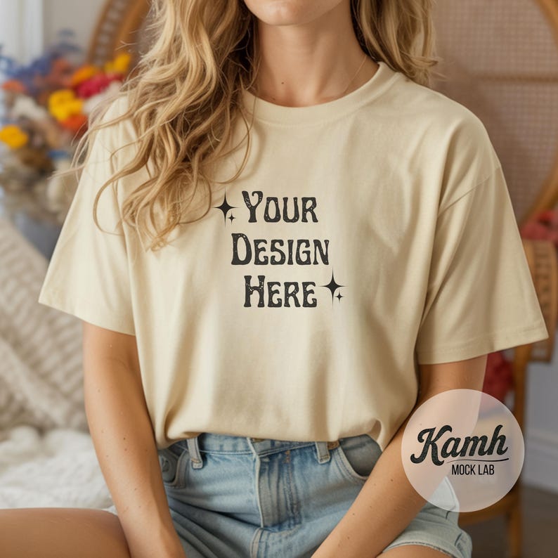 May include: Cream-colored t-shirt with the text "YOUR DESIGN HERE" in a black, vintage-style font. The shirt is a short-sleeved, crew neck style. The model is wearing blue denim shorts.