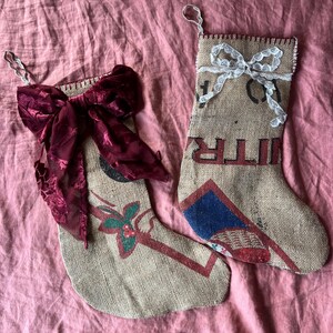 May include: Two burlap Christmas stockings with different embellishments. One stocking has a large burgundy bow and the other has a white lace bow.