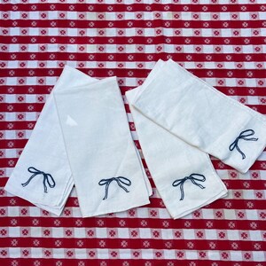 以下が含まれることがあります： 青い刺繍のリボンが付いた白いリネンナプキンが4枚。ナプキンは折り畳まれ、赤と白のチェック柄のテーブルクロスの上に置かれています。