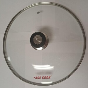 May include: A circular, transparent glass pot lid featuring a silver-toned metal rim and a central black knob with a silver metallic insert. The glass surface displays the red text ACE COOK with a small logo symbol positioned above the brand name.