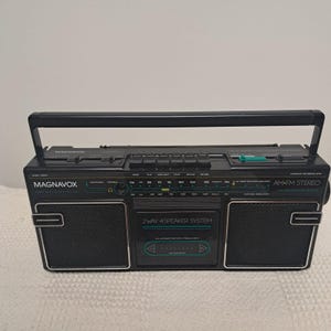 May include: A black Magnavox portable AM/FM stereo cassette recorder featuring a top handle, two front-facing speakers, and a central cassette deck compartment. The control panel includes buttons for playback functions and a radio frequency tuning dial.