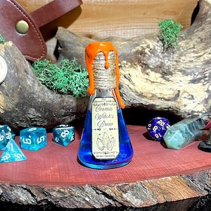 May include: A still life arrangement featuring a blue potion bottle with orange wax, dice, a figurine, and a purple crystal ball. The bottle is labeled "Cosmic Witch's Brew". The items are arranged on a wooden surface.