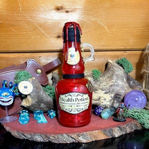 May include: A red glass bottle labeled "Health Potion" with a gold chain and handle. The bottle has a black top and a label that reads "Drink to restore life essence. Do not consume." The bottle is on a wooden surface.