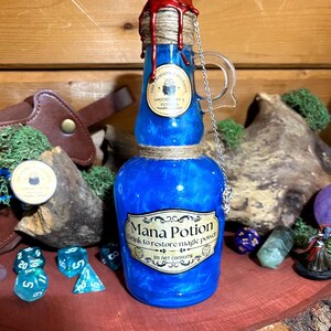 May include: A blue glass potion bottle with a red wax seal and a label that reads "Mana Potion". The bottle is decorated with twine and a silver chain. The text on the label says "Drink to restore magic power".