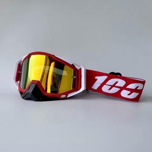 May include: White and red motocross goggles with a reflective yellow lens. The goggles have a red strap with the white text "100" printed on it. The goggles have a black nose guard.