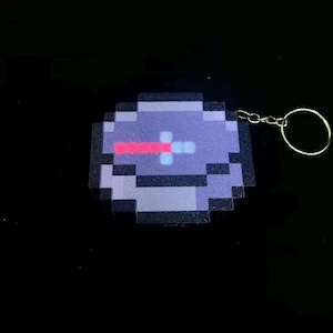 Op de afbeelding: Pixelige kompas sleutelhanger in tinten paars, grijs en rood. De sleutelhanger heeft een pixel art ontwerp van een kompas, met een goudkleurige sleutelring bevestigd. Het ontwerp doet denken aan retro videogames.