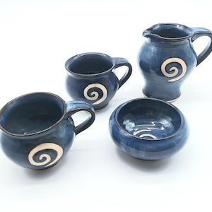 May include: A set of four handmade ceramic pieces in a deep blue glaze. The set includes two mugs, a small pitcher, and a small bowl. Each piece features a white spiral design. The pieces are arranged on a white surface.