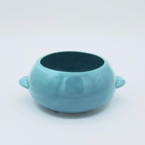 May include: A round, light blue ceramic bowl with a dark interior. The bowl has two small handles on either side. The glossy finish reflects light, and the overall design is simple and modern. This bowl is suitable for serving or display.