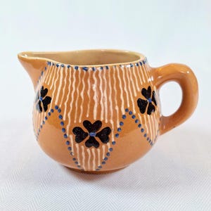 May include: A small, brown ceramic pitcher with a white and blue floral design. The pitcher has a rounded body and a handle.