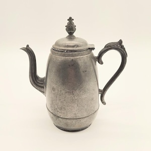 May include: A vintage pewter teapot with a curved spout and ornate handle. The lid is topped with a decorative finial. The teapot has a bulbous body and a dark, aged appearance, suggesting it is an antique.