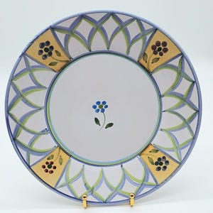 May include: A white ceramic plate with a hand-painted floral design. The plate features a blue and green geometric border, gold accents, and a central blue flower. The plate is displayed on a gold stand.