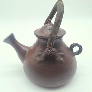May include: A brown ceramic teapot with a wooden handle and lid. The teapot has a spout and a small loop handle on the side. The handle is made of a curved piece of wood.