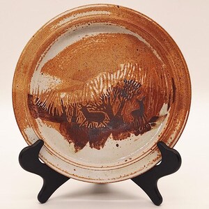 May include: A handmade ceramic plate with a rustic design. The plate features a brown and white color scheme with a landscape scene of trees and deer. The plate is displayed on a black stand.