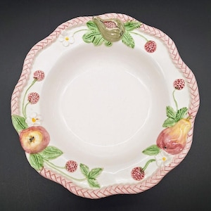 May include: A white ceramic serving bowl with a scalloped edge and a braided pink border. The bowl is decorated with raised relief fruit and floral designs, including figs, pears, apples, raspberries, and small white flowers.